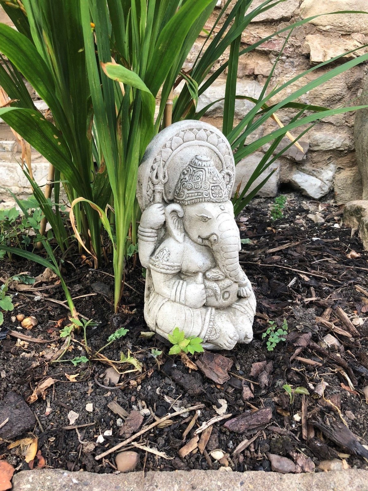 Ganesh Garden Ornament - Small Stone nestled among green leaves and soil. Durable limestone statue for garden decoration.