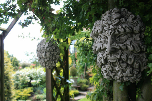 Garden Wall Plaques - Greenman Greenwoman in solid stone finish mounted on wooden trellis, surrounded by lush greenery for outdoor decor.
