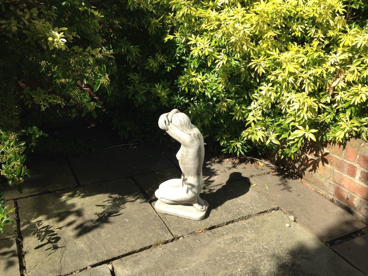 Stone garden statue of nude woman kneeling, placed on paved patio with sunlight and shadow. Decorative outdoor art piece.
