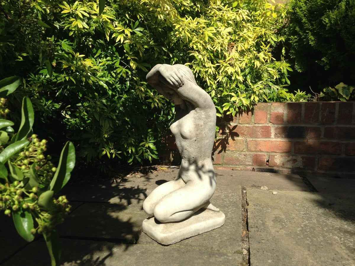 Limestone nude lady statue kneeling with arms overhead, positioned on stone patio. Elegant garden ornament for modern outdoor decor.