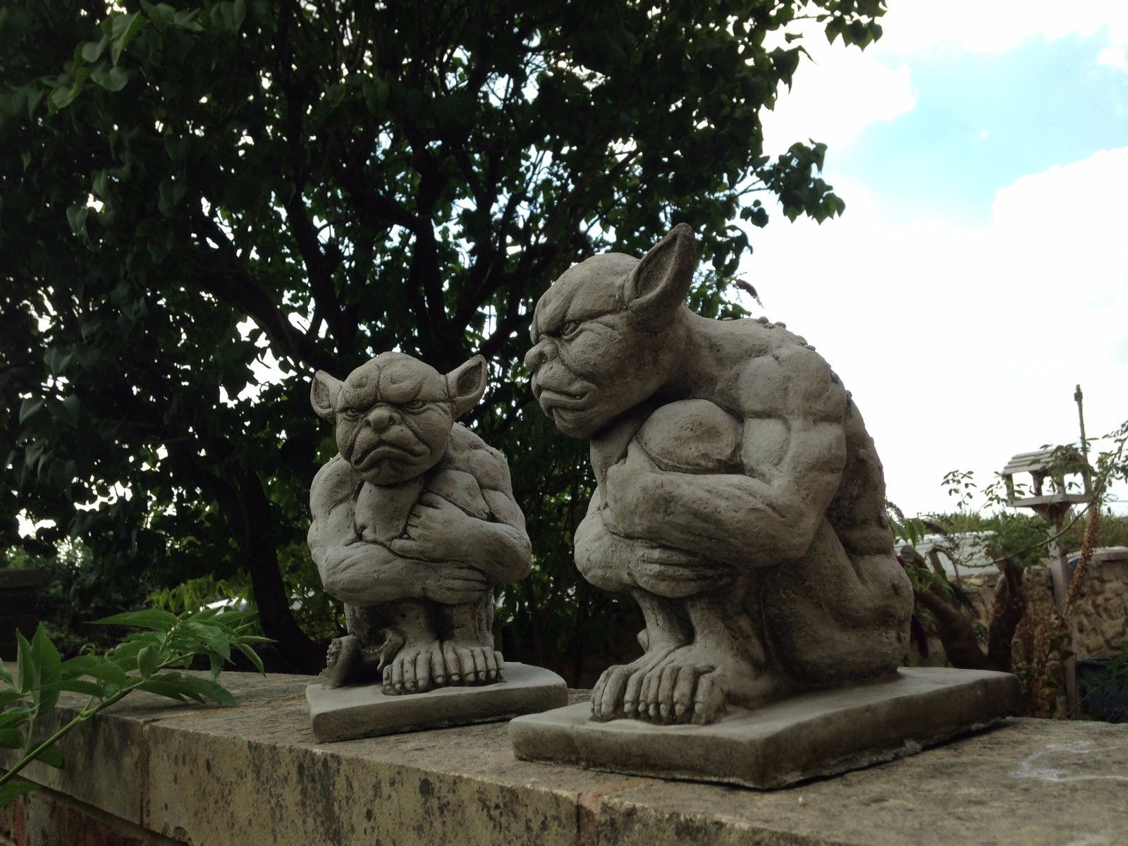 Close-up of Grumpy Stone Gargoyle pair on patio wall. Handcrafted limestone statues for weatherproof outdoor garden decor.