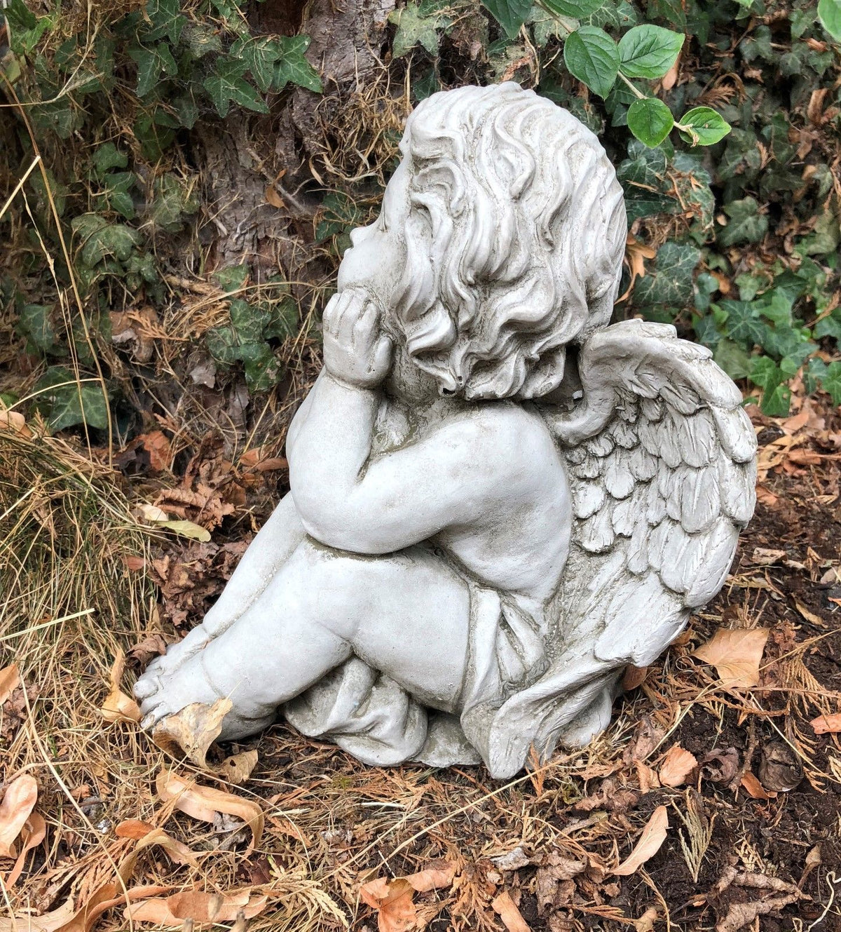 Stone winged baby angel garden ornament sitting with hands on cheeks, detailed limestone sculpture for outdoor decor in flowerbeds or patios.