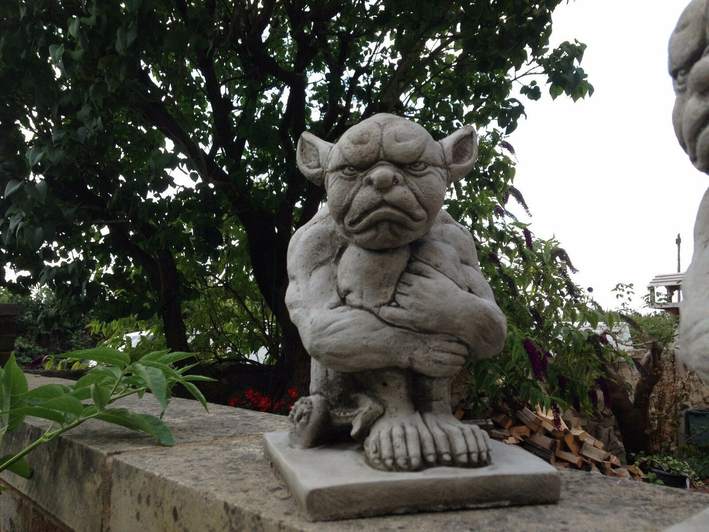 Single Grumpy Gargoyle Statue on stone ledge with tree backdrop. Ideal for gothic garden decor and lawn ornamentation.