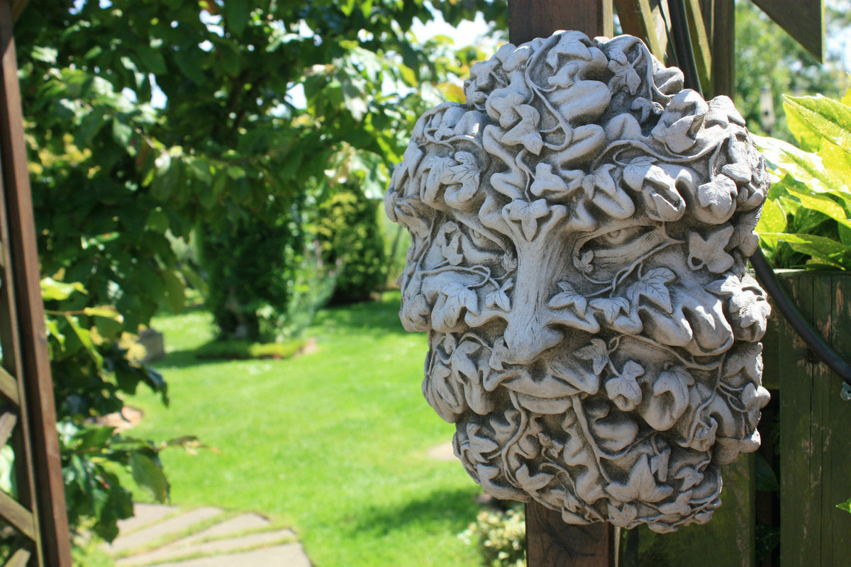 Greenman wall plaque in solid stone finish with intricate leaf design, mounted on wooden post in a vibrant garden setting.