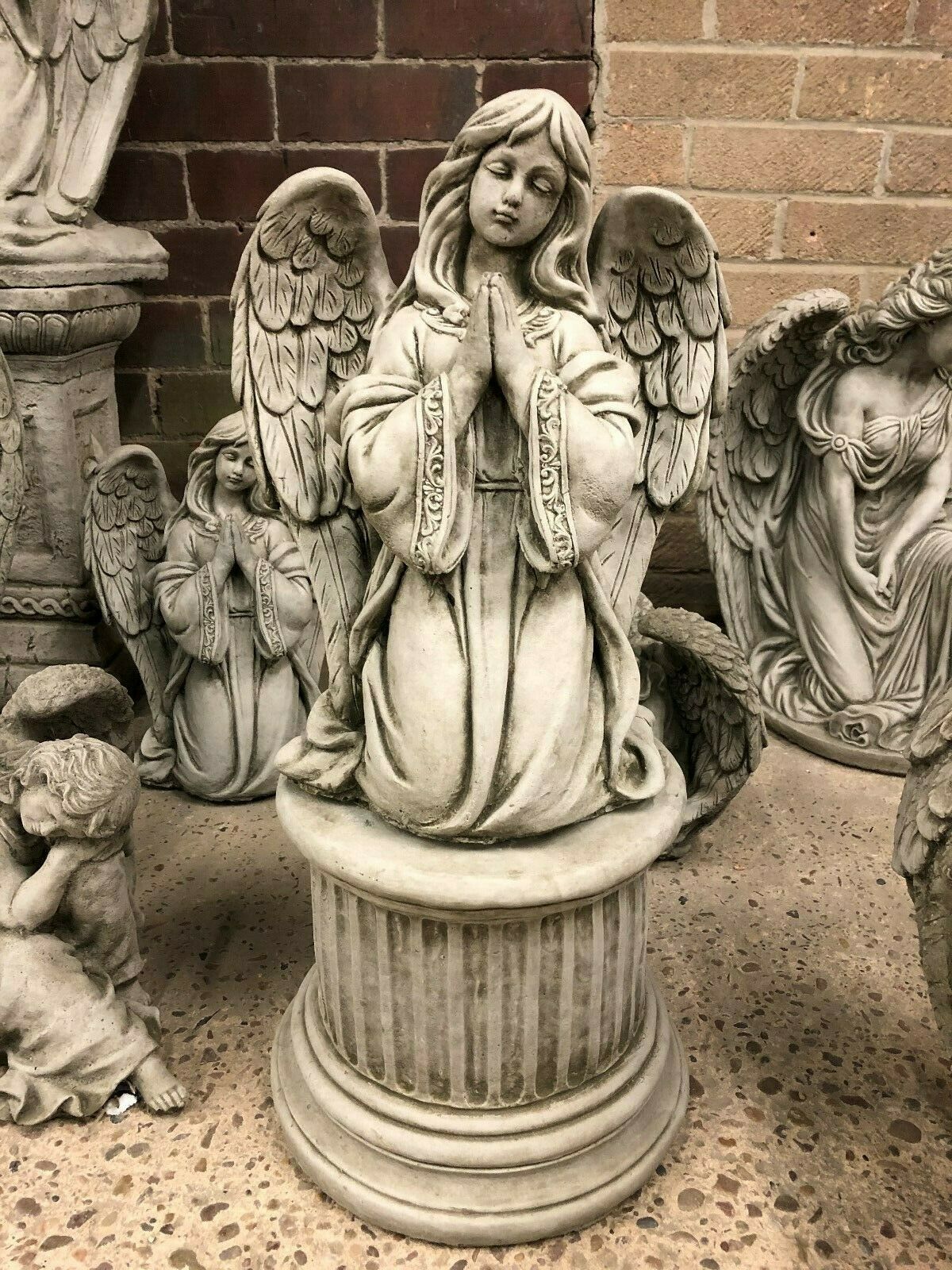 Durable Stone Praying Angel Statue with antique finish, kneeling on a fluted pedestal. Ideal for elegant garden decor and outdoor spiritual ambiance.