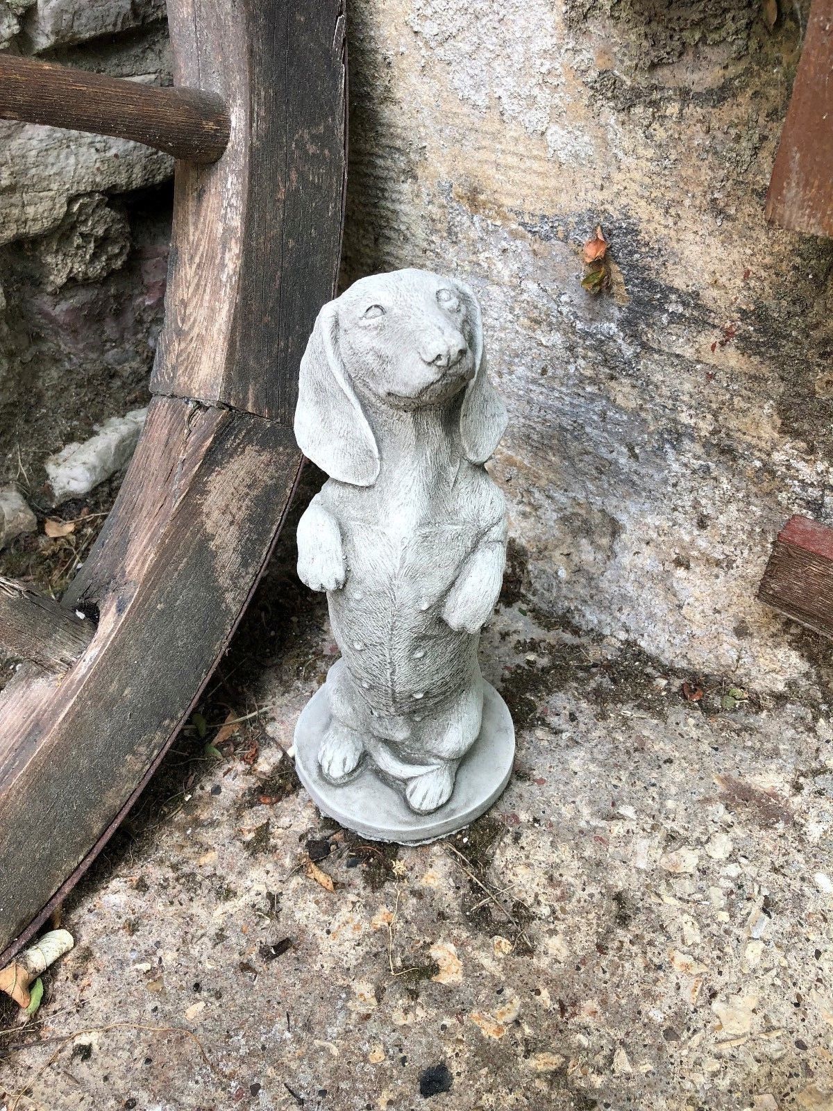 Handcrafted limestone dachshund sculpture placed by a rustic wall. Great decorative garden animal ornament for patios and flower borders.