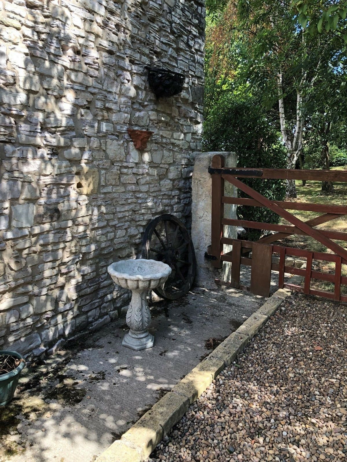 Antique finish stone bird bath with leaf design placed in rustic garden corner near stone wall and wooden gate.