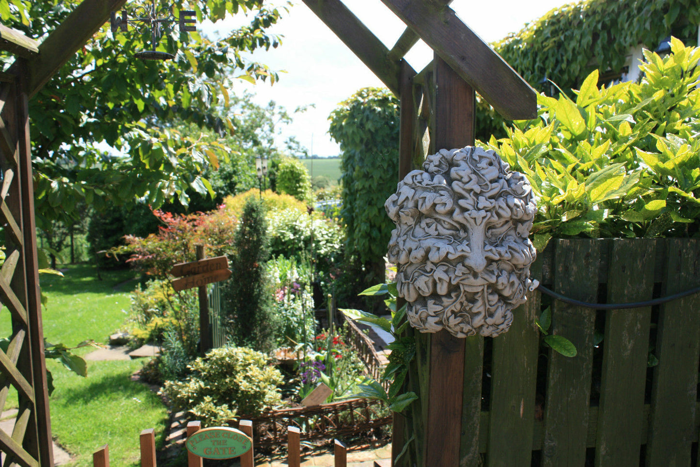 Greenman garden wall plaque in solid limestone with carved foliage face, mounted on a rustic wooden fence in a landscaped garden.