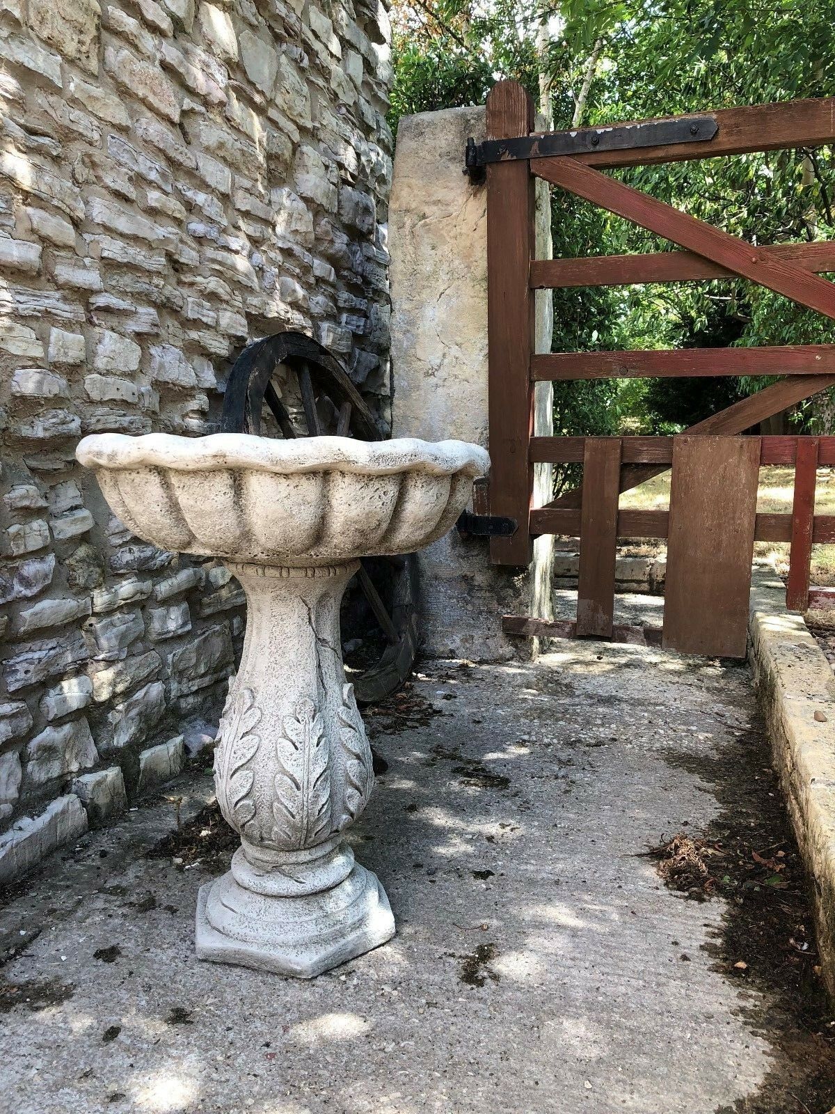Elegant garden bird bath with carved leaf pedestal and weathered stone texture, positioned on patio near wooden gate.