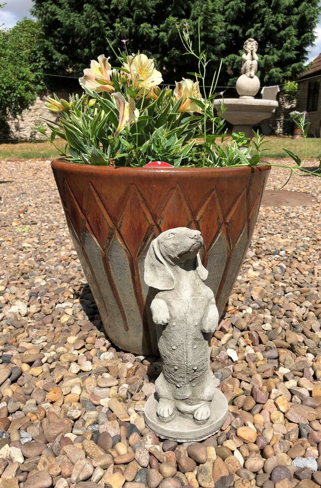 Stone dachshund sculpture standing beside a flower pot on gravel. Ideal garden decor piece with a sturdy and weatherproof stone finish.