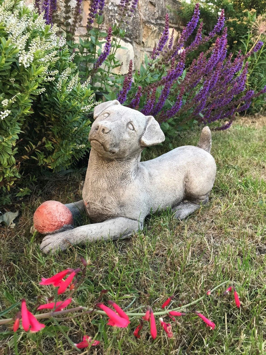 Jack Russell Dog Ornament - Stone Garden Decor featuring a lifelike dog statue holding a red ball, set on grass with purple flowers in the background.