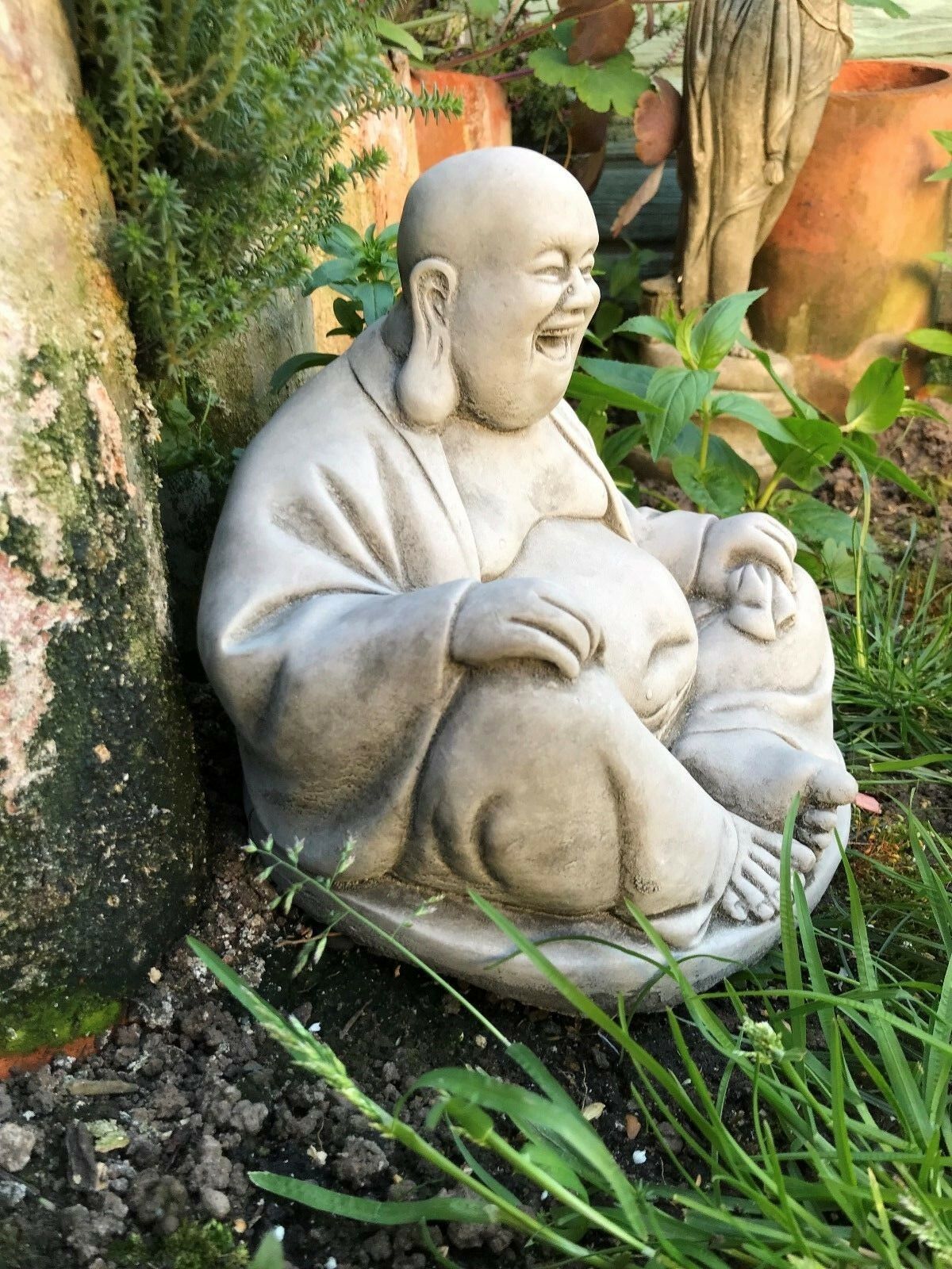 Angled view of Stone Laughing Buddha Ornament placed among garden plants, showcasing its serene and joyful design for zen spaces.
