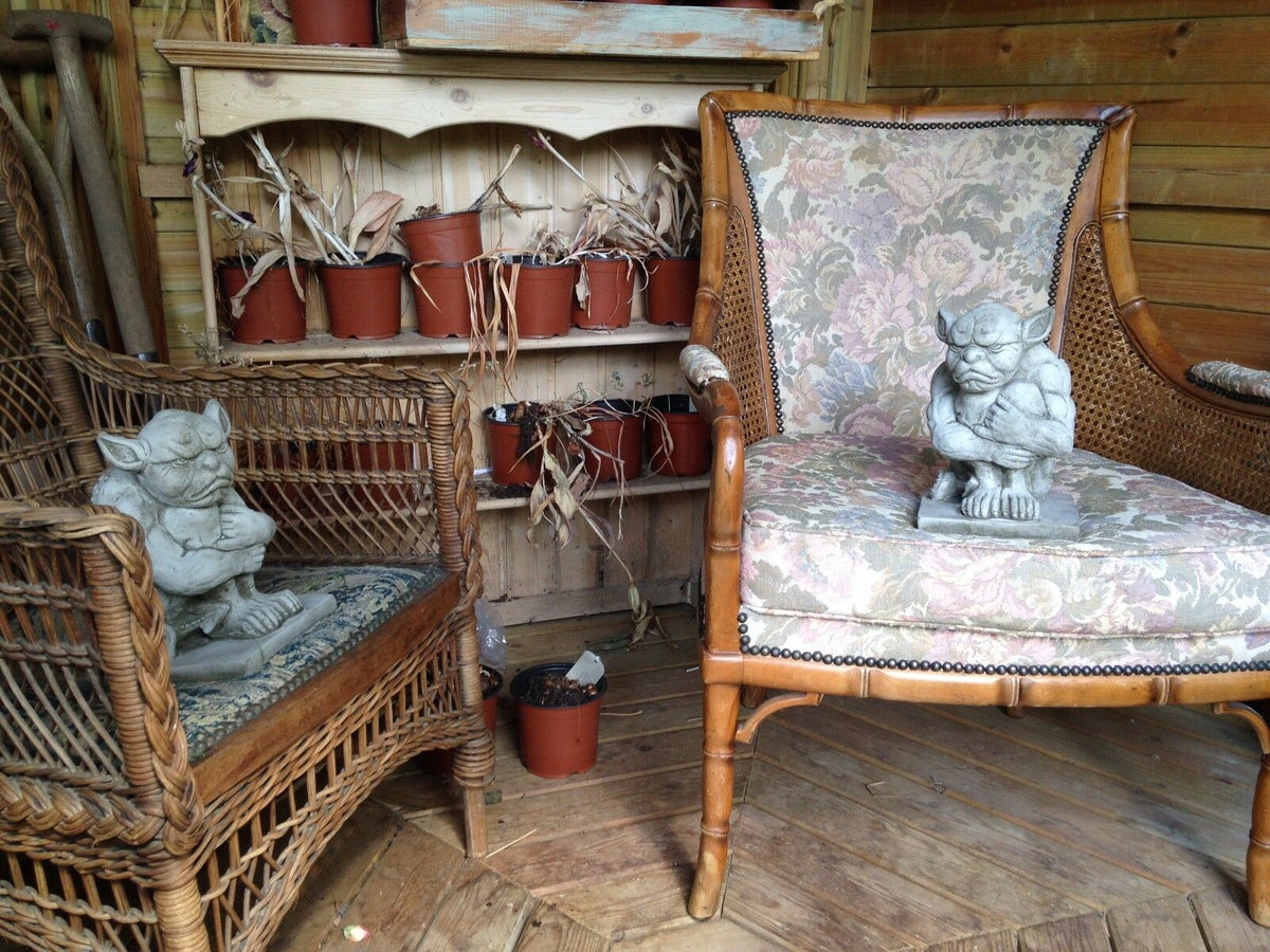 Pair of Grumpy Gargoyle Statues seated on wicker chairs in rustic room. Unique gothic garden ornaments for indoor display.