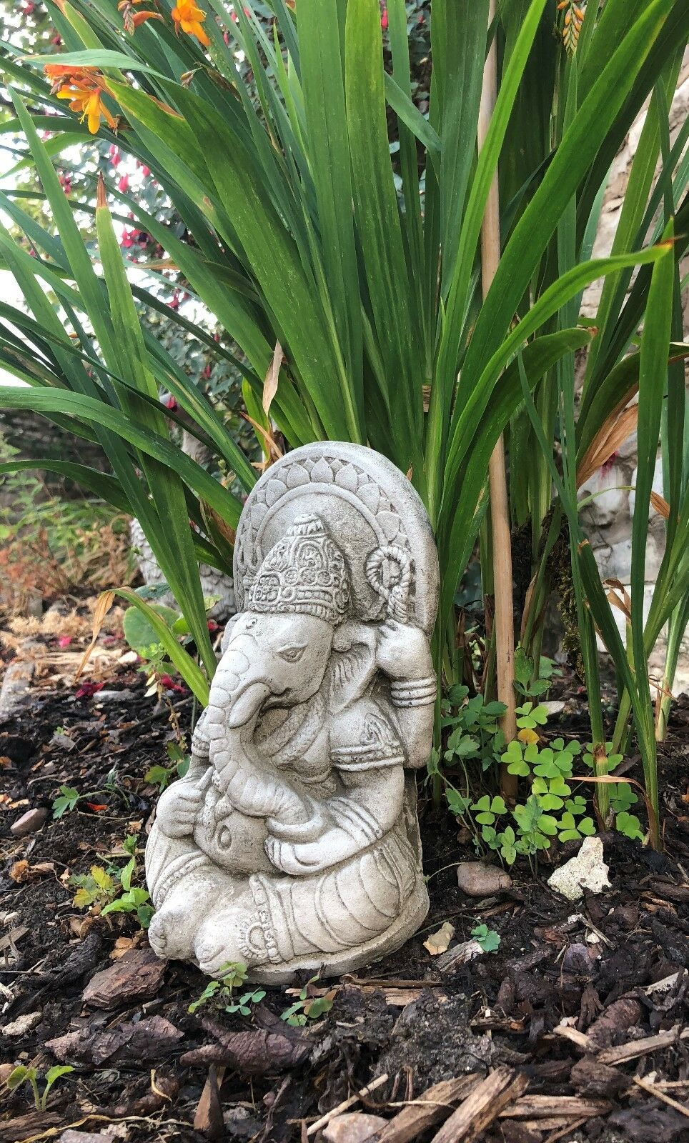 Ganesh Garden Ornament - Small Stone statue beside tall green plants. Durable and spiritual garden ornament for outdoor spaces.