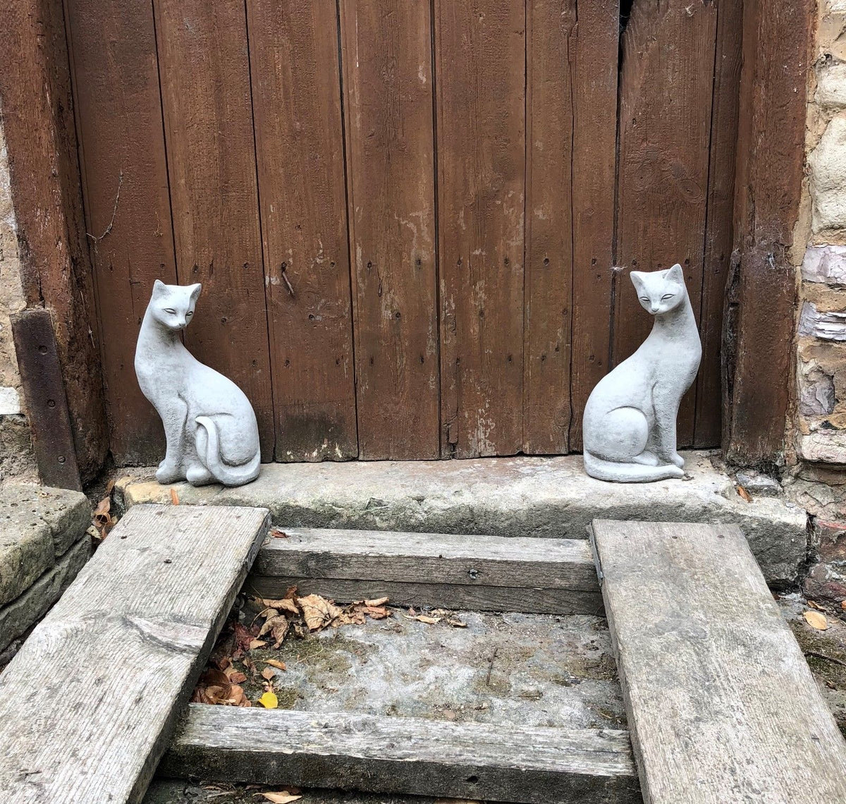 Pair of stone Siamese cat statues placed symmetrically by a rustic wooden door, enhancing garden entryways with elegant charm.