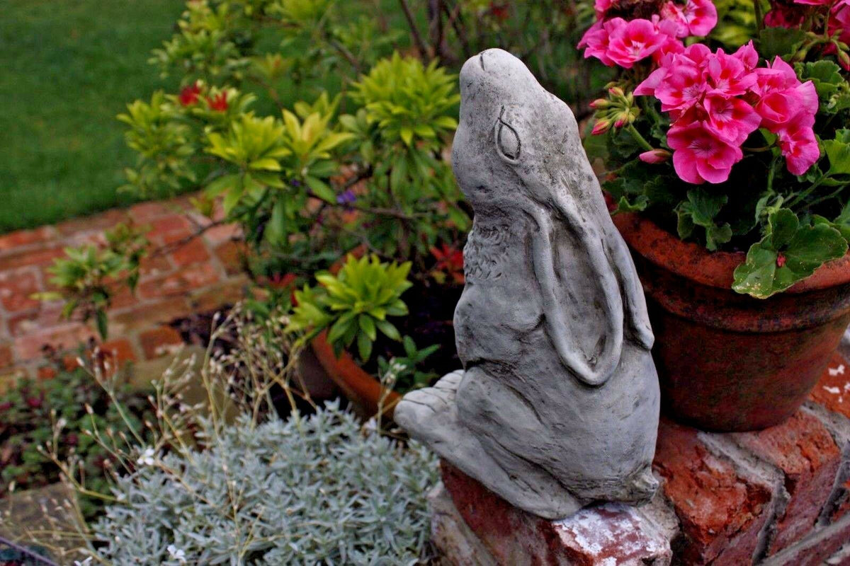 Moongazing Hare Stone Statue placed in a vibrant garden with pink flowers and greenery, ideal for outdoor garden decor and lawn ornamentation.