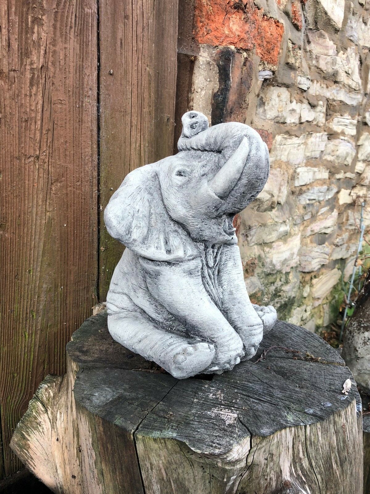 Stone baby elephant statue with raised trunk on a wooden stump, a decorative garden feature for animal lovers and outdoor spaces.