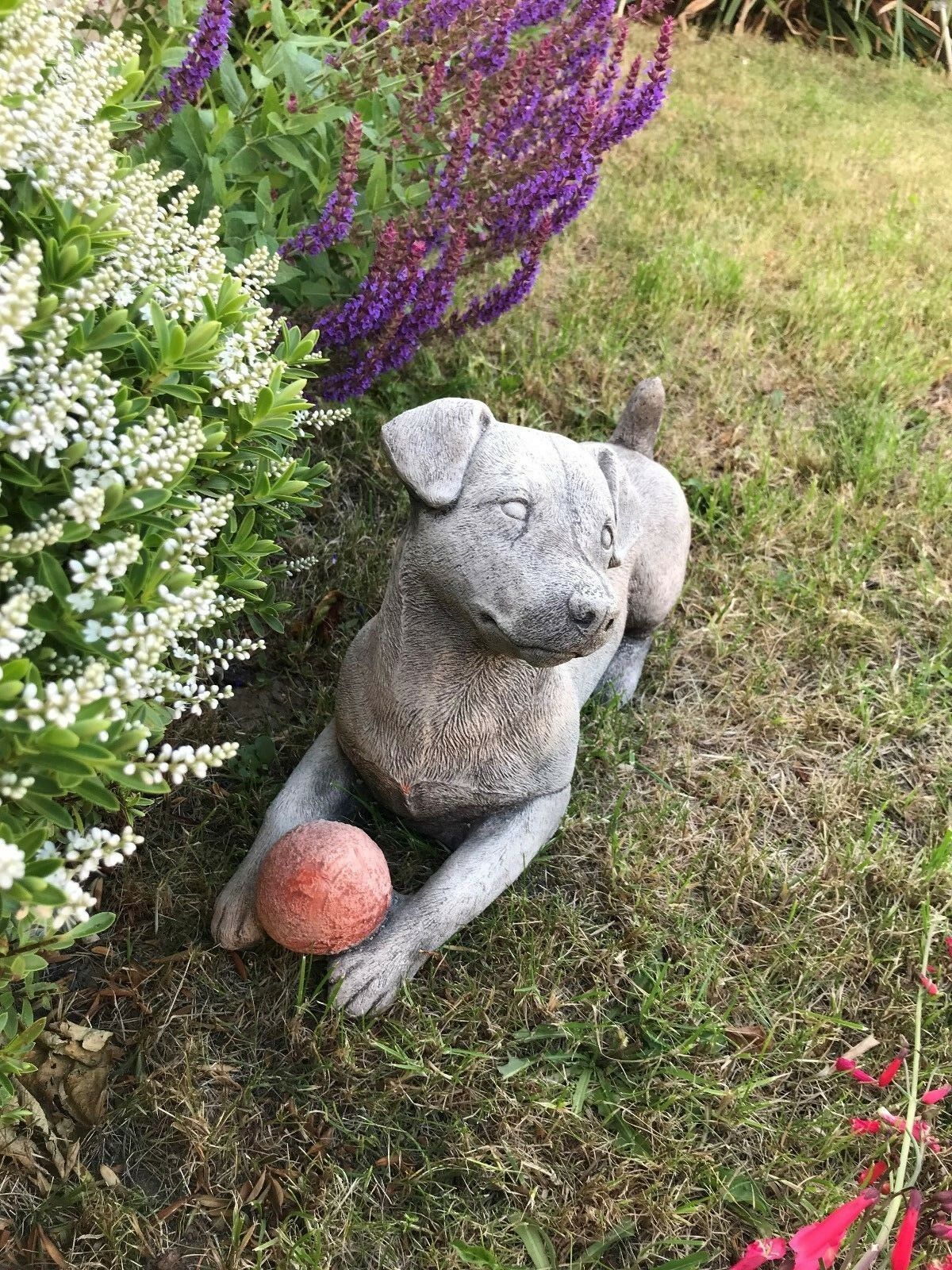 Stone Jack Russell Dog Ornament with red ball, positioned on garden grass with blooming purple and white flowers in the background.