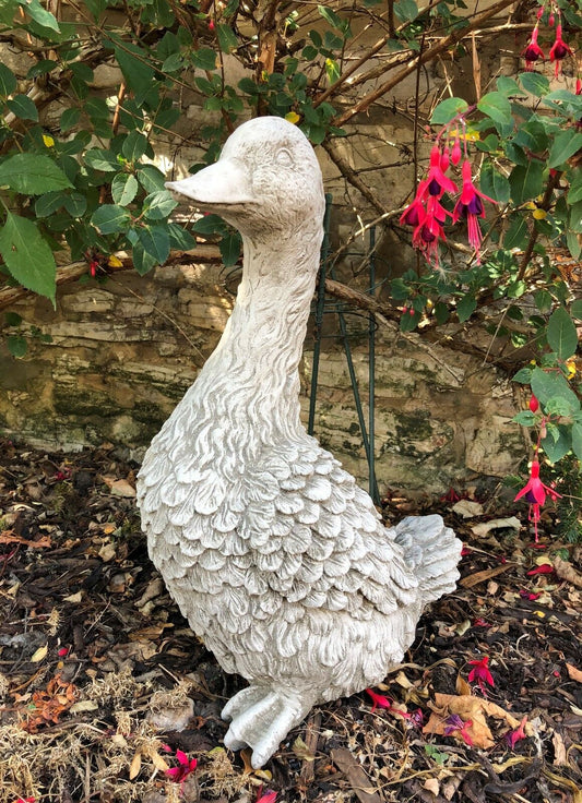 Goose Garden Statue - Stone Goose Decor placed in a flower bed with red fuchsia blooms. Durable ornament for outdoor garden decoration.