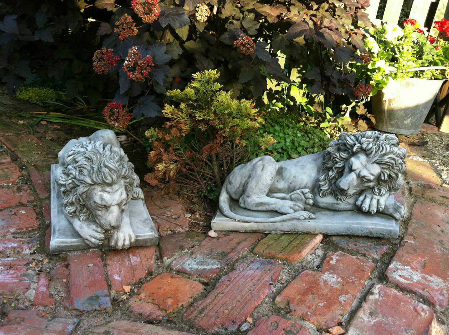 Pair of stone lion garden statues lying on brick path near foliage, offering classic and durable decor for patios, gates, or flowerbeds.