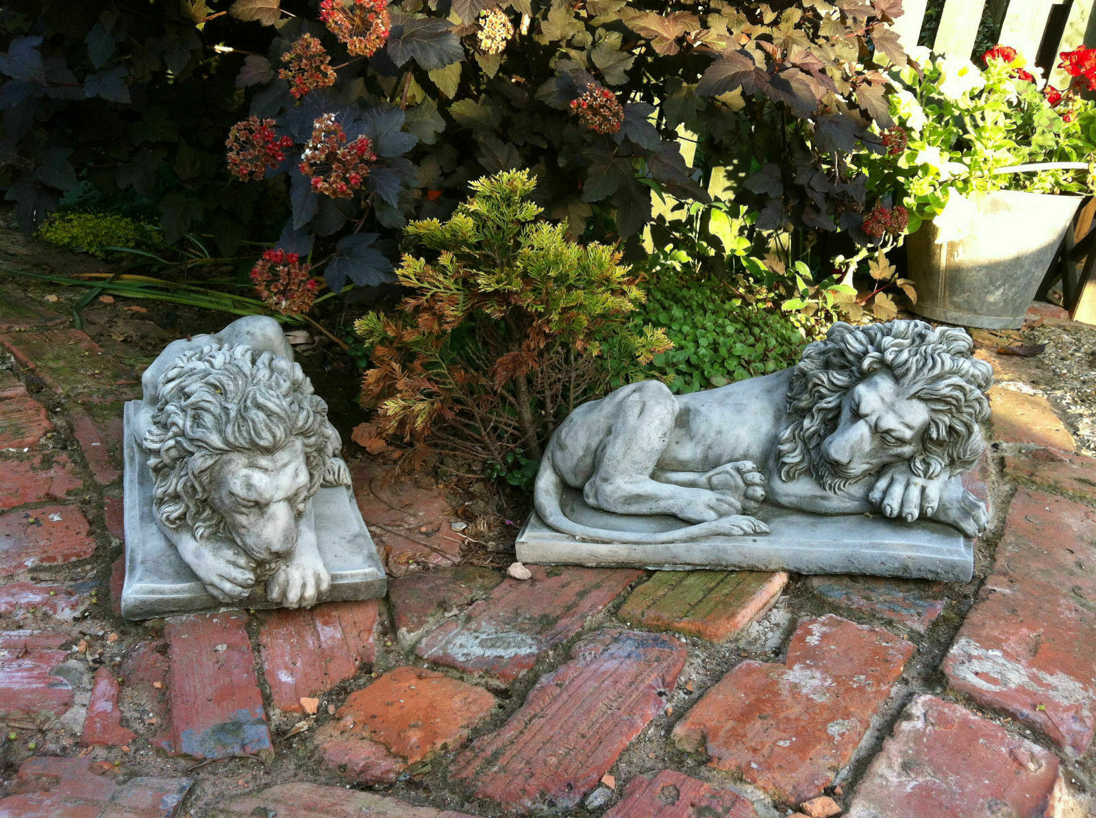 Pair of stone lion garden statues lying on brick path near foliage, offering classic and durable decor for patios, gates, or flowerbeds.