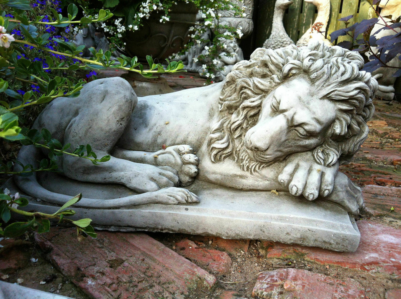 Elegant stone lion statue with head resting on paw, surrounded by garden plants, ideal for timeless outdoor decor and pillar-top display.