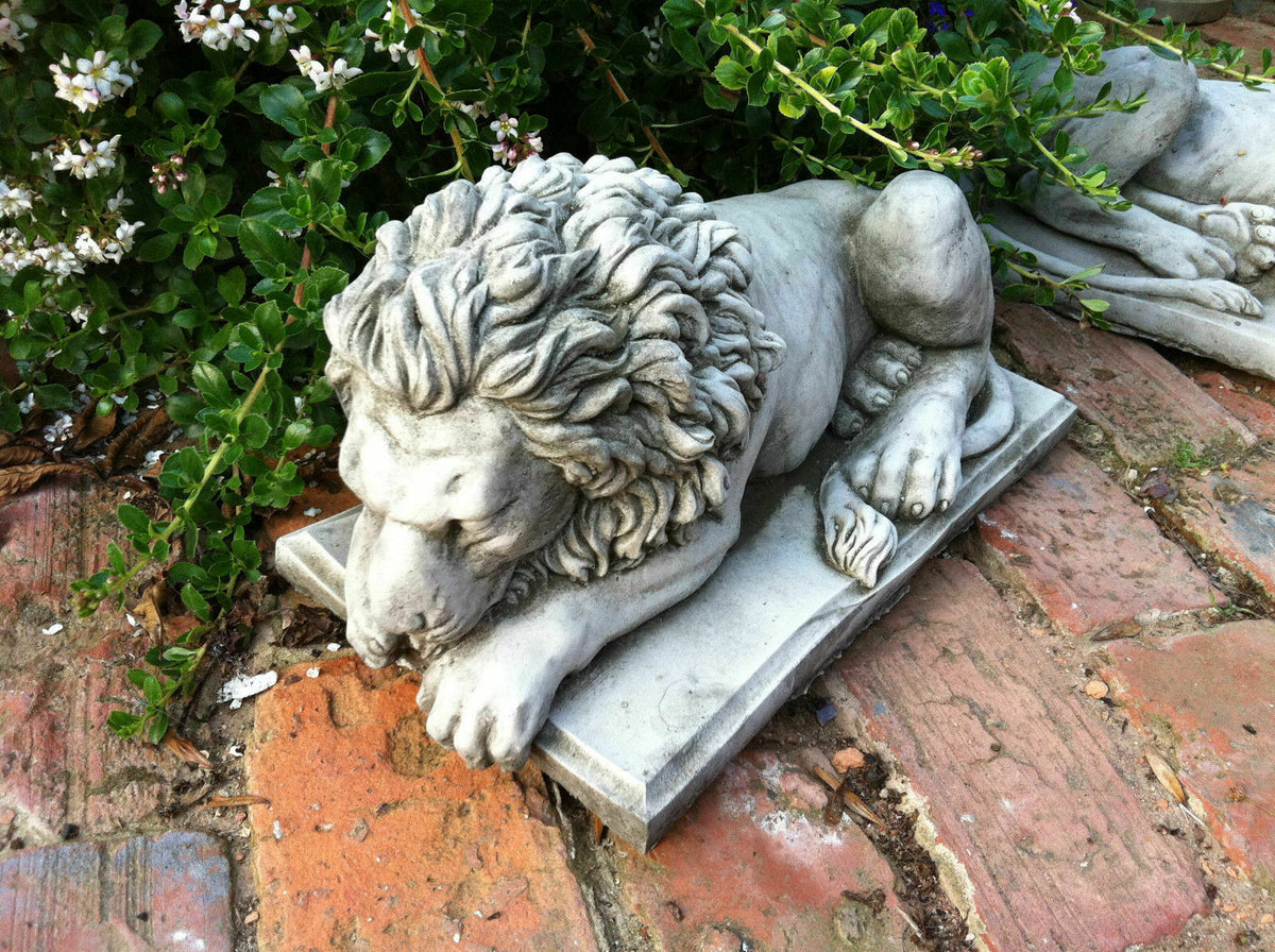 Detailed stone lion statue lying peacefully on a base, placed on a garden path with greenery, perfect for classic lawn ornamentation.