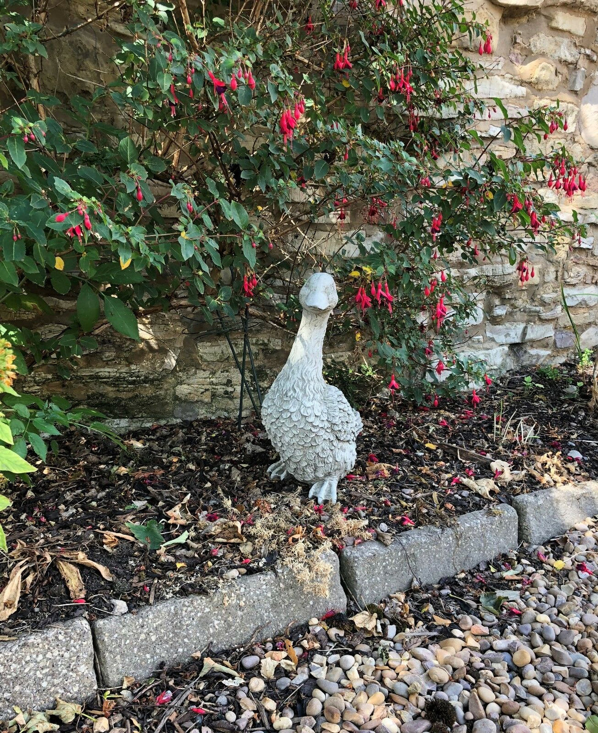 Stone Goose Garden Statue nestled among red flowers and mulch. A charming outdoor ornament for home garden decoration.