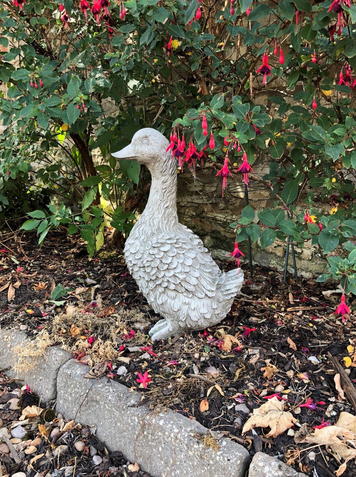 Stone Goose Garden Statue surrounded by vibrant red flowers. A decorative lawn sculpture perfect for patios and flowerbeds.