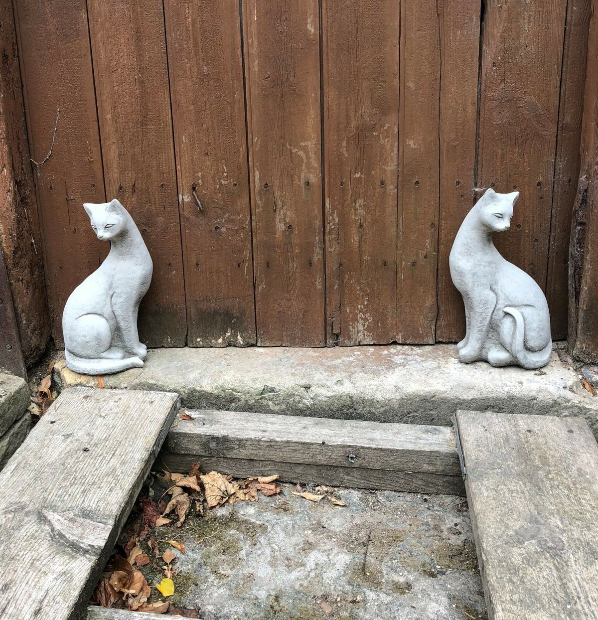 Weatherproof stone Siamese cat statues flanking a wooden garden door, adding symmetry and style to outdoor landscaping.