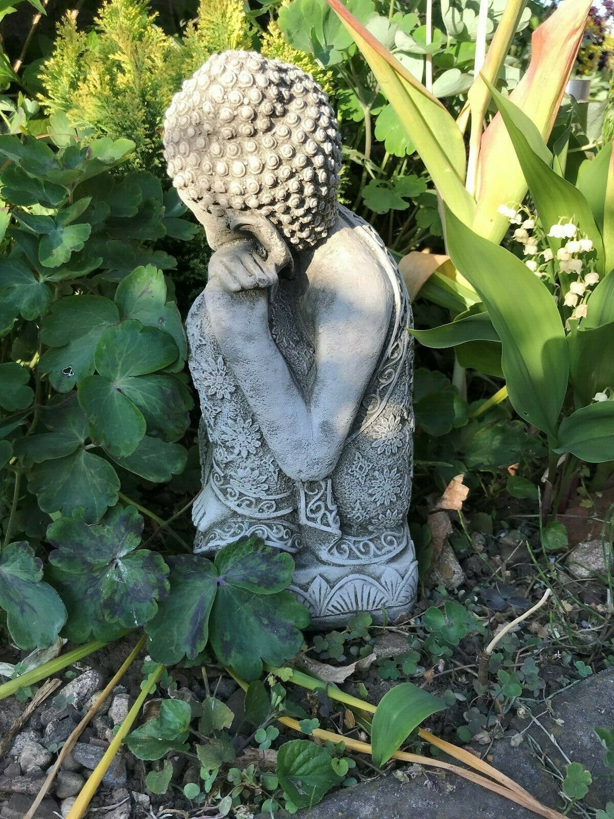 Angled view of limestone Sleeping Buddha statue with detailed robe design, nestled among garden plants for a peaceful outdoor ornament.