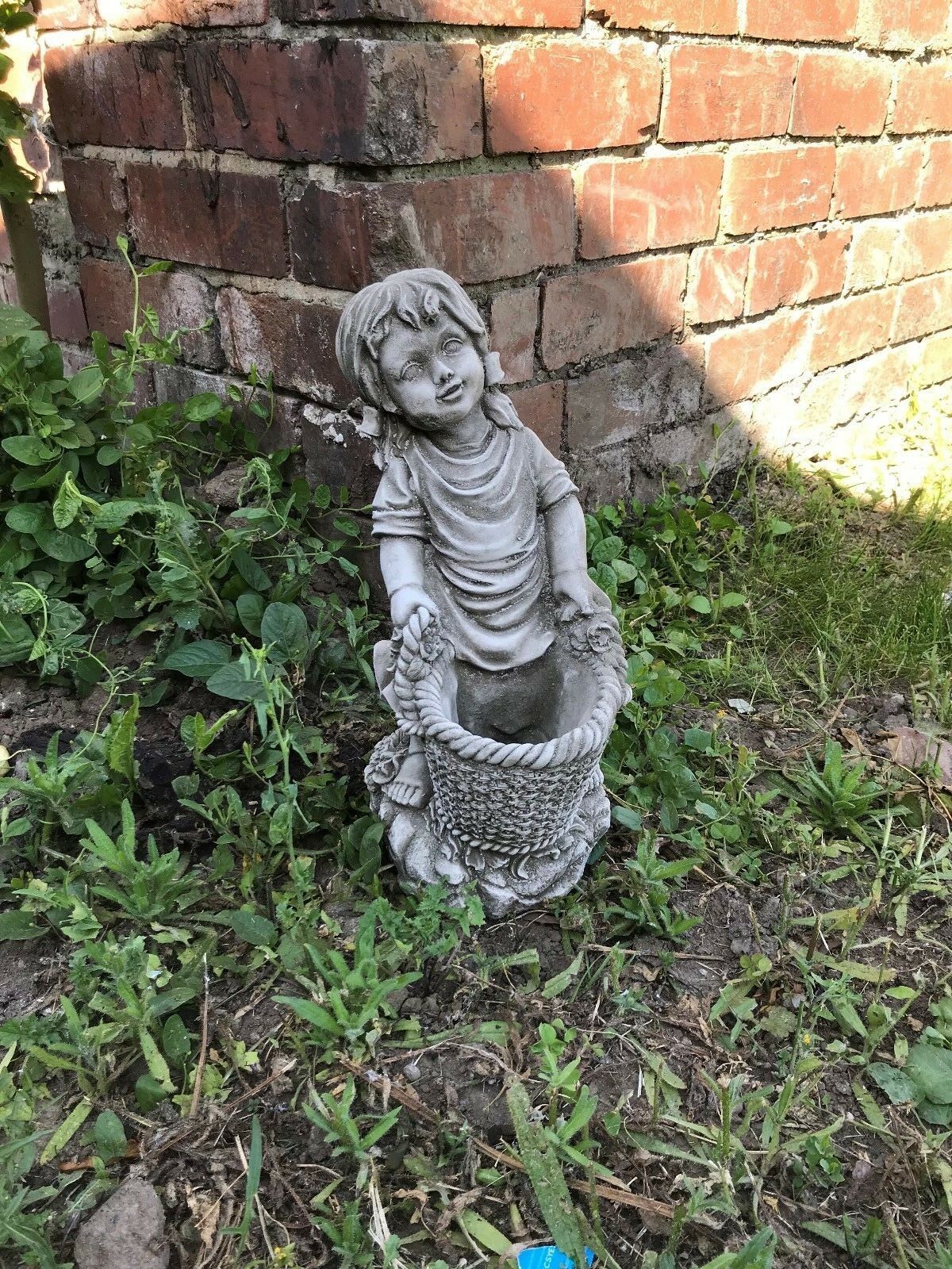 Stone Basket Girl Garden Planter Ornament placed on garden soil near a brick wall, ideal for rustic outdoor decor.