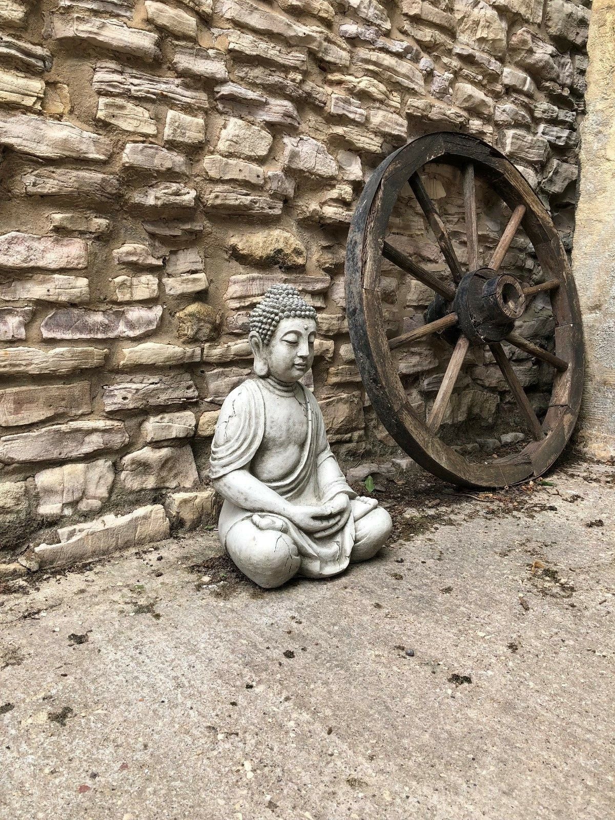 Full view of Stone Sitting Buddha Statue beside vintage wooden wheel, enhancing rustic garden charm and outdoor spiritual decor.