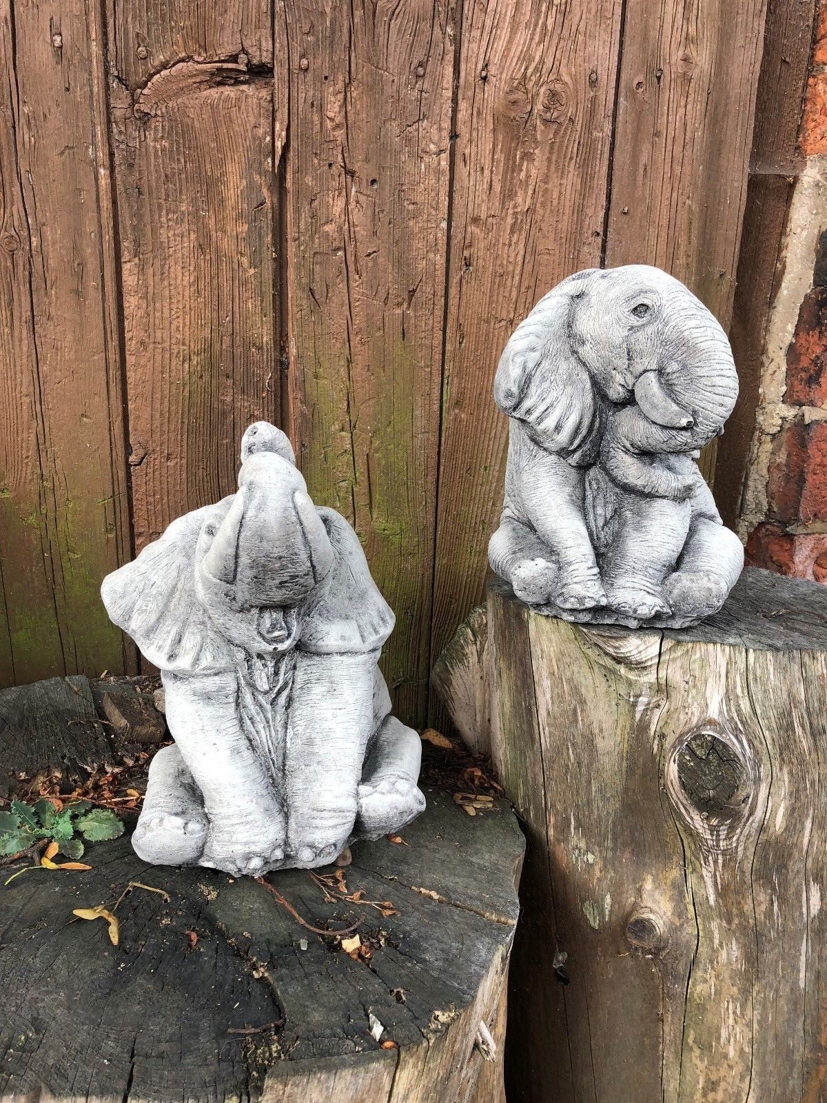 Two baby elephant garden ornaments in grey stone finish, posed on tree stumps for rustic outdoor decor in home gardens or patios.