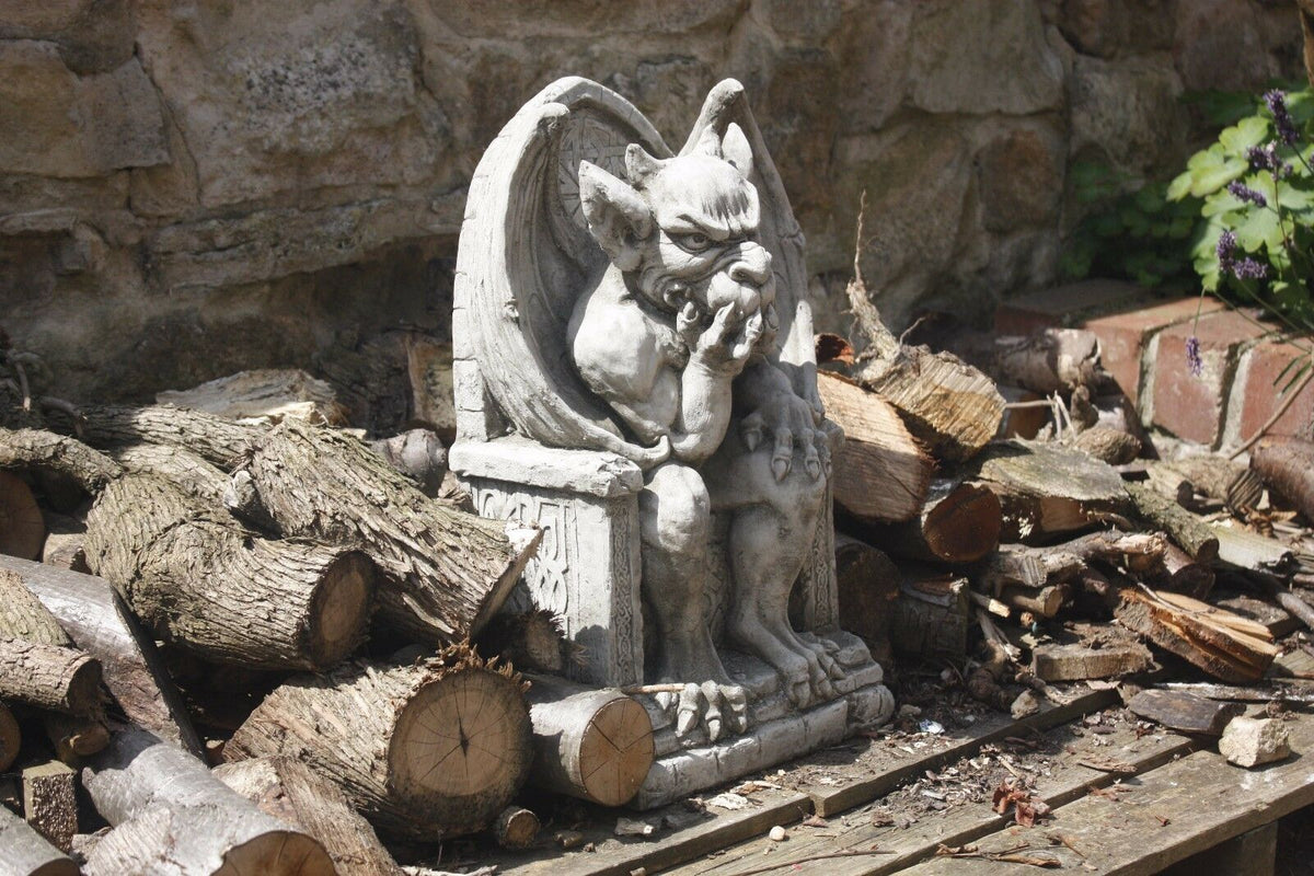 Side outdoor view of King Gargoyle Stone Statue - Weather Resistant Garden Art surrounded by logs, showcasing its rugged and decorative garden use.