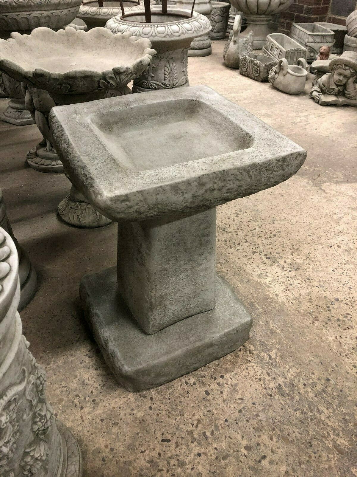 Angled view of stone bird bath with thick square edges and pedestal. Antique grey finish complements traditional garden settings.