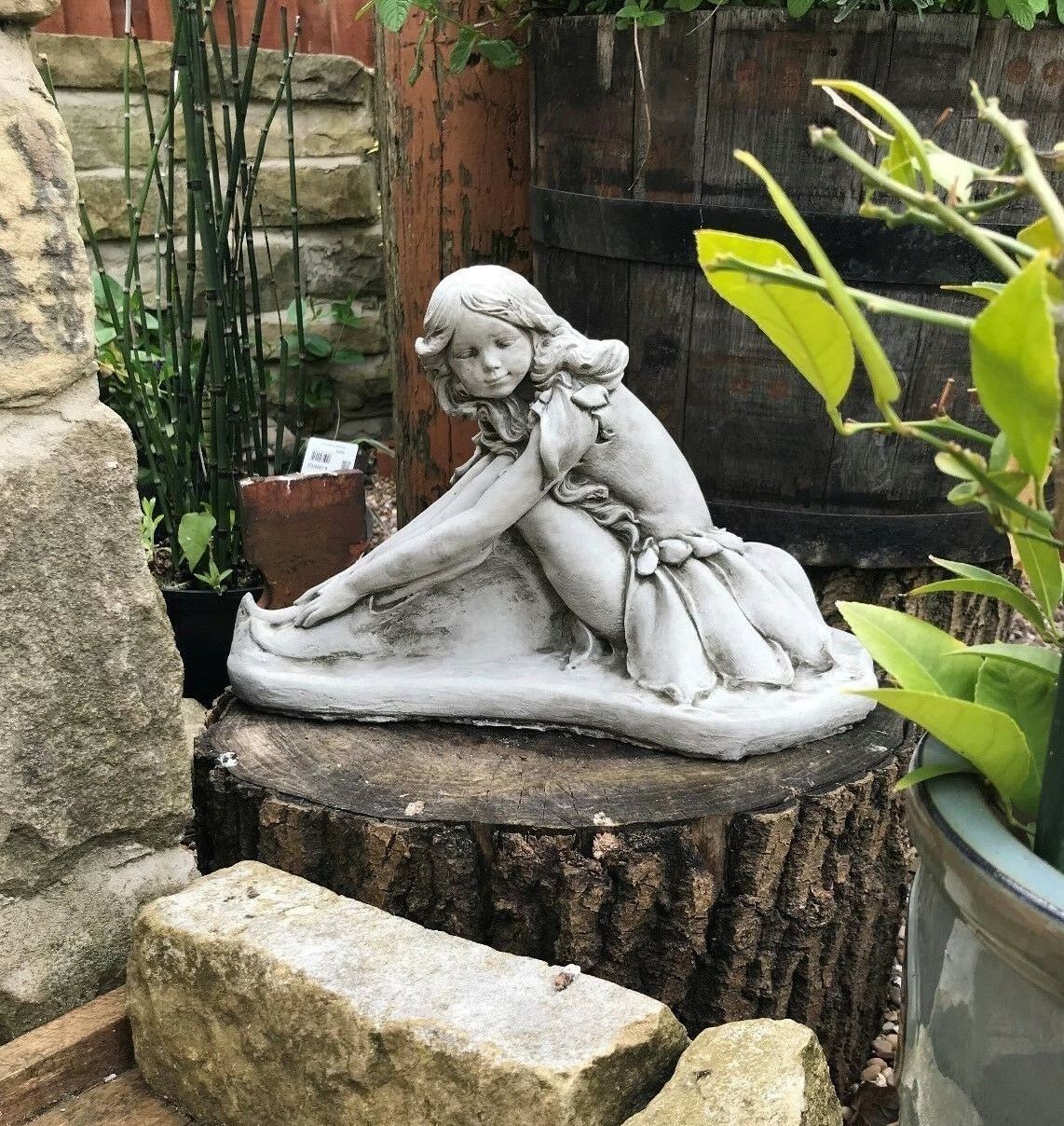 Handmade stone fairy sculpture placed among garden plants and wooden barrels, enhancing outdoor charm with its detailed craftsmanship.