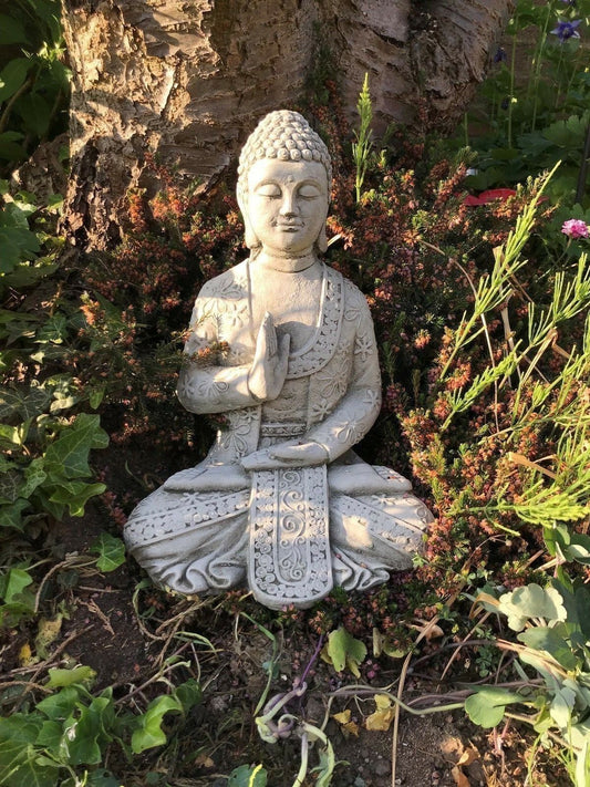 Stone Buddha Garden Statue in meditative pose with floral detailing, placed under a tree among garden plants for outdoor serenity décor.