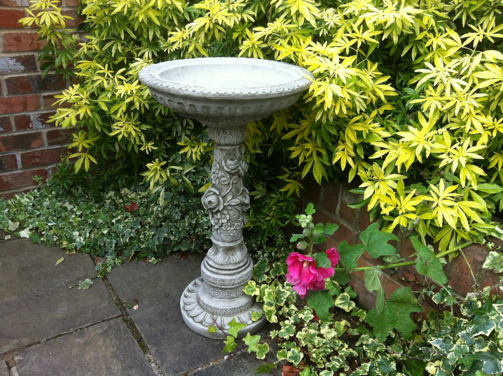 Bird Bath - Stone Flower Petal Design in a garden corner with lush yellow-green foliage. Elegant limestone finish adds charm to outdoor decor.