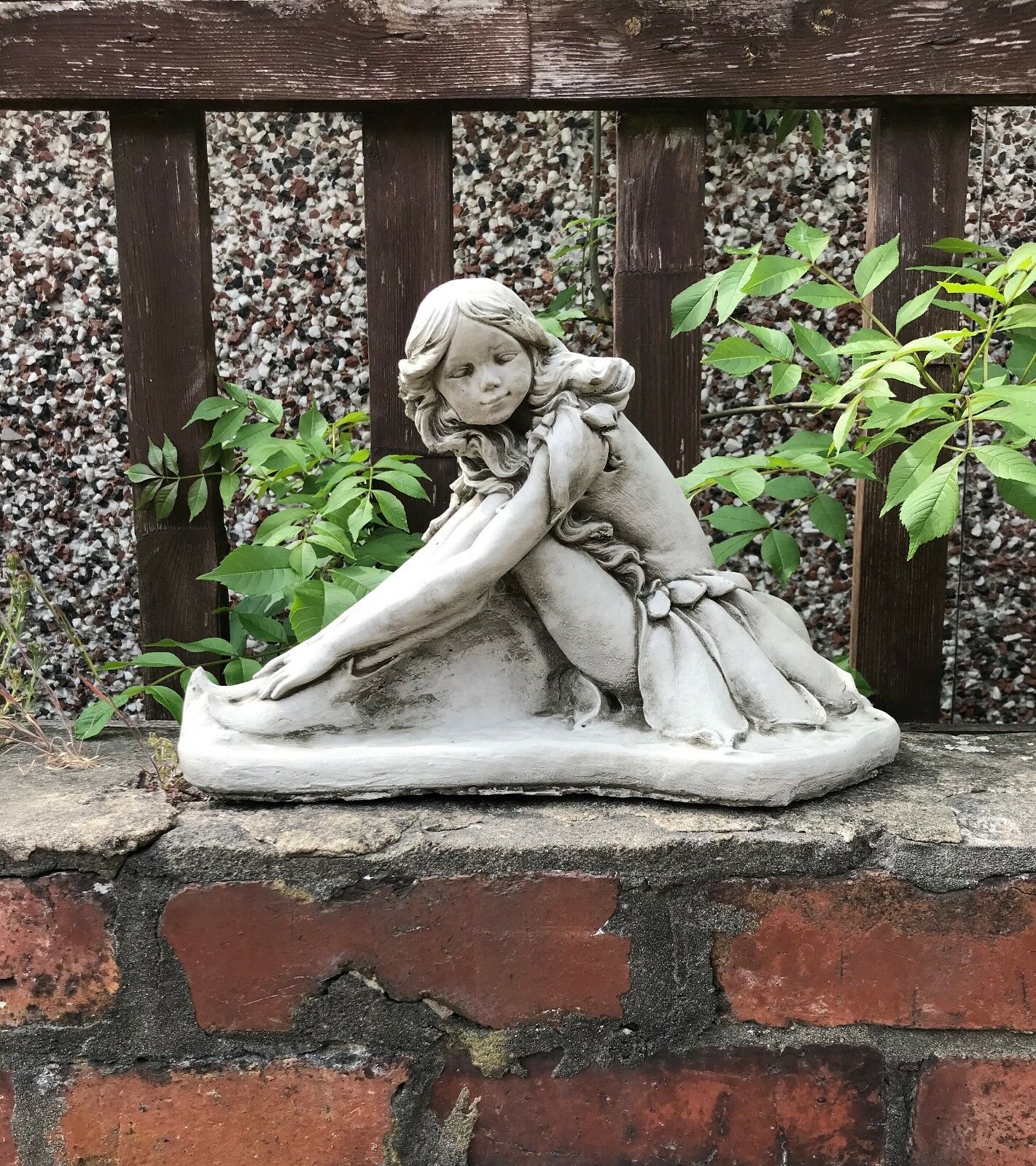 Stone fairy garden ornament displayed on a brick wall with wooden fence backdrop, adding a magical touch to outdoor garden decor.