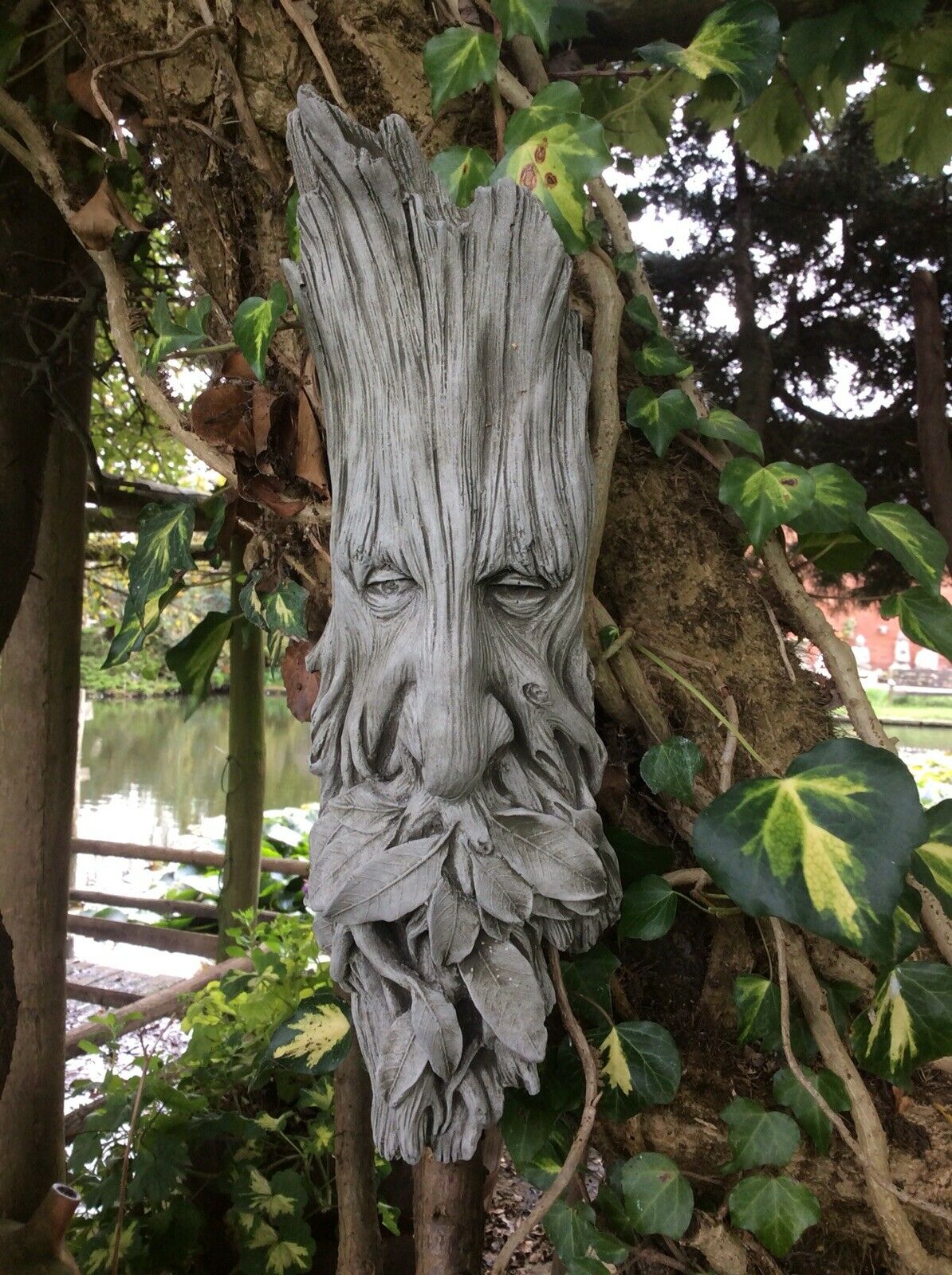 Mythical Greenman wall plaque in stone leaf finish, mounted on a tree with lush garden background, ideal for outdoor decoration.