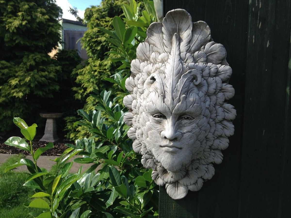 Stone Feather Greenman Wall Plaque displayed outdoors with vibrant foliage. Weather-resistant limestone plaque for garden or patio decor.