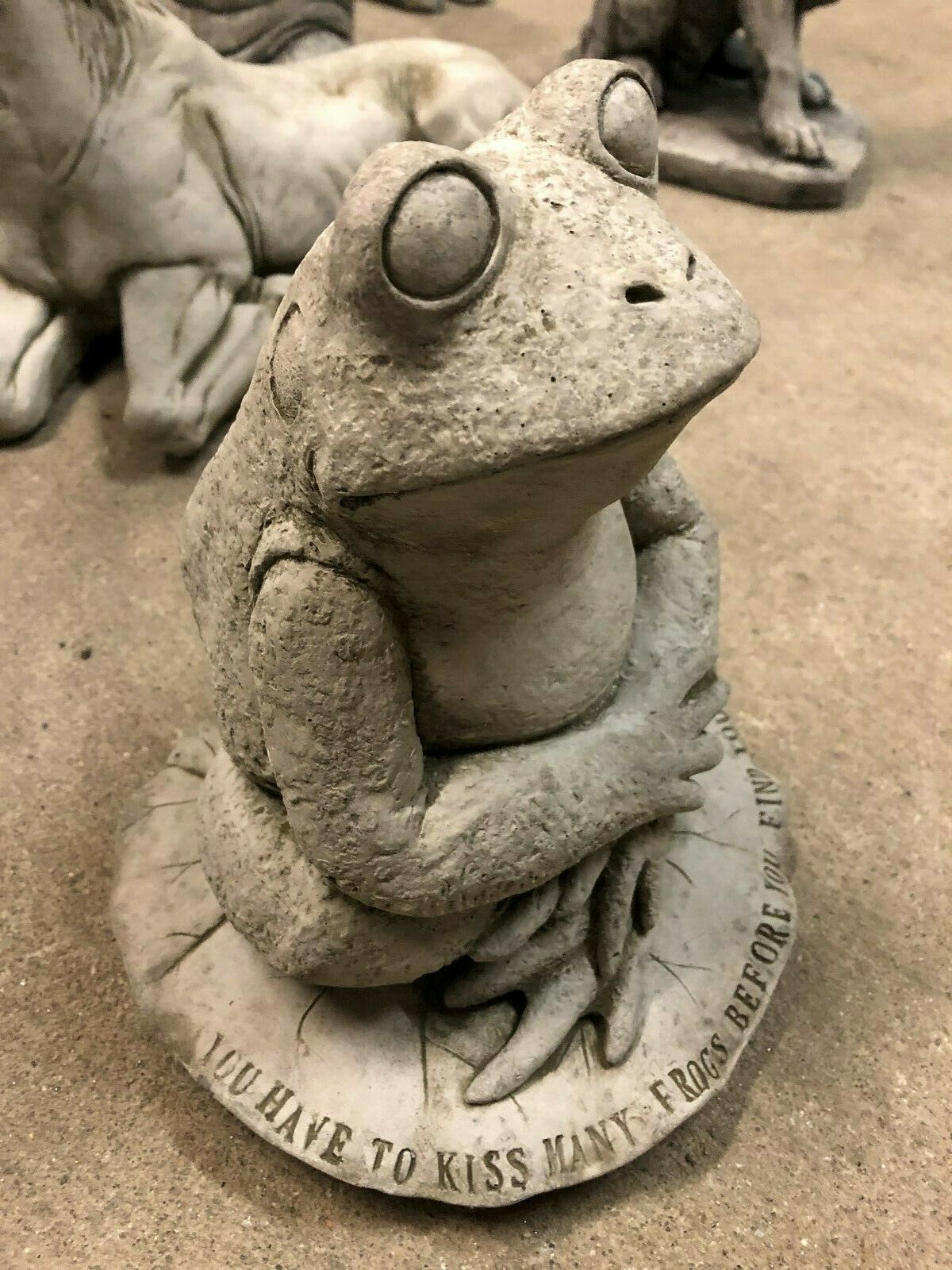 Close-up of the frog garden ornament with expressive eyes and folded arms, sitting on a lilypad with a witty engraved message around the base.