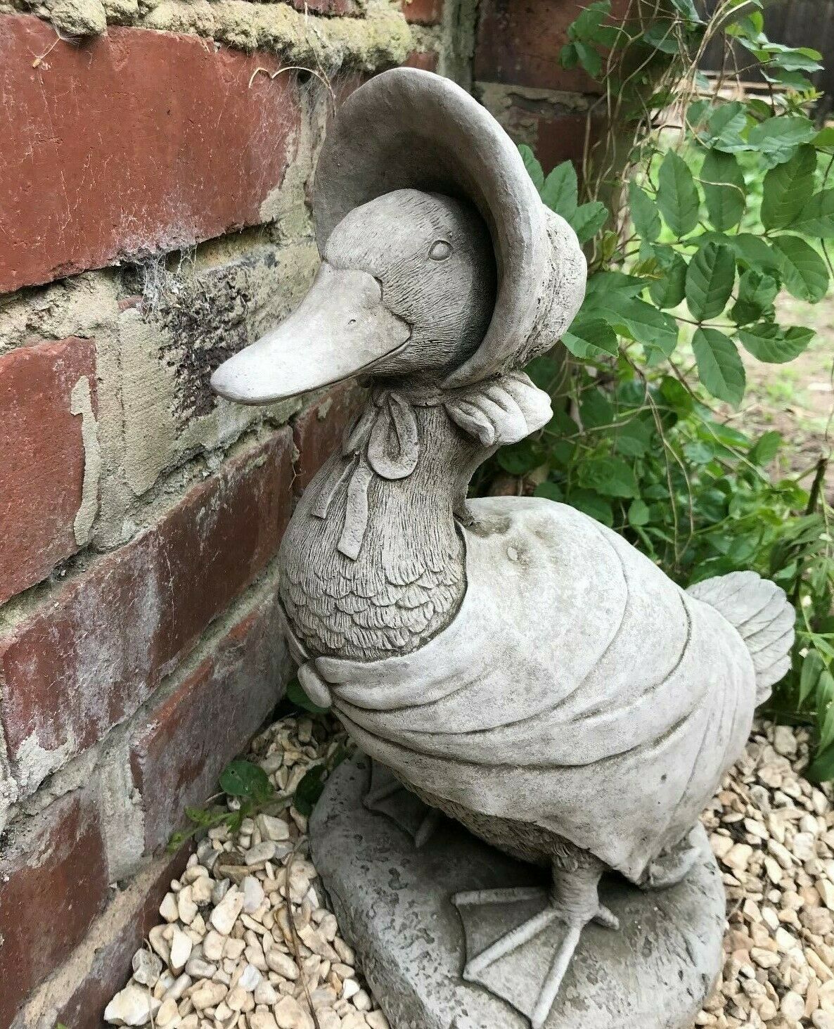 Jemima Duck garden ornament made from durable stone, placed on gravel beside a brick wall. Ideal for outdoor lawn or flowerbed decor.