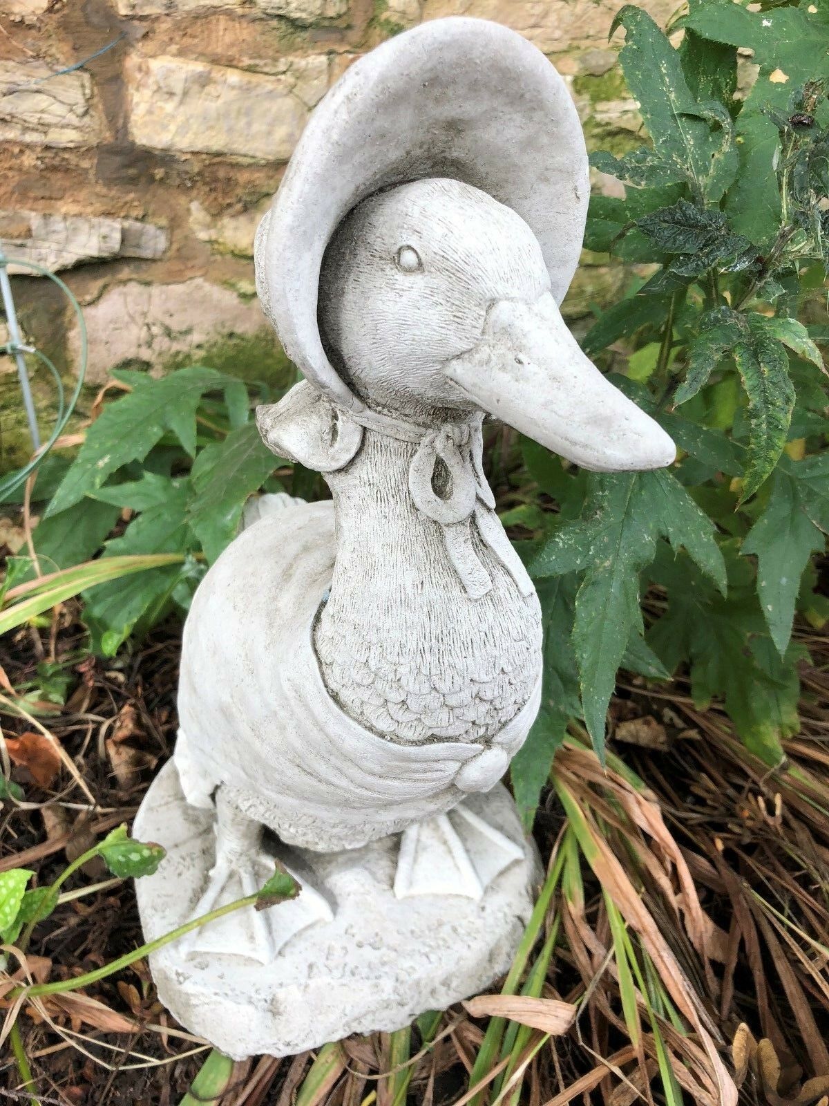 Charming Jemima Duck stone ornament in bonnet and shawl, set among garden plants. Perfect for enhancing garden sculpture collections.