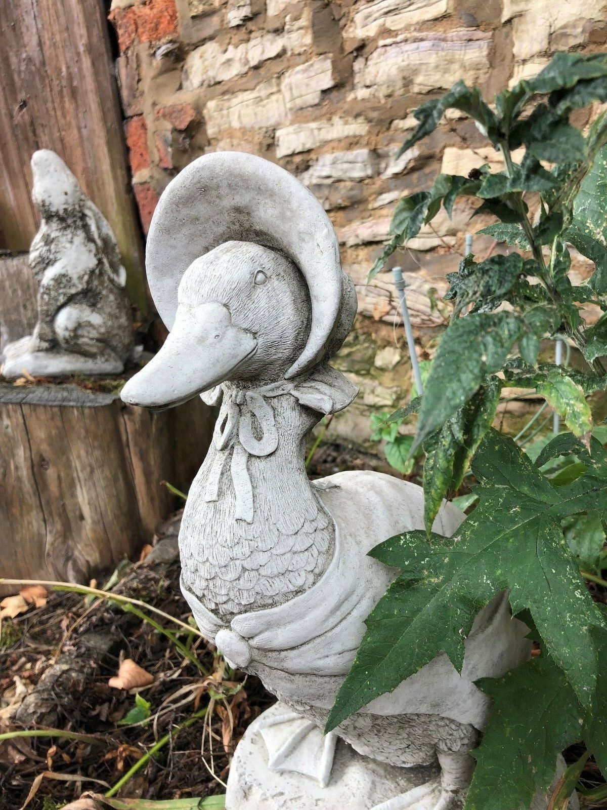 Detailed stone duck statue with bonnet, surrounded by greenery and rustic textures. A whimsical addition to garden animal ornaments.