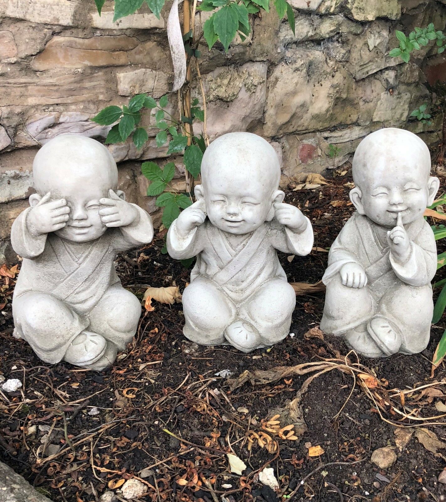 Baby Monk Ornaments - See, Hear, Speak No Evil Set in stone finish, displayed in a garden setting with rustic stone wall background.