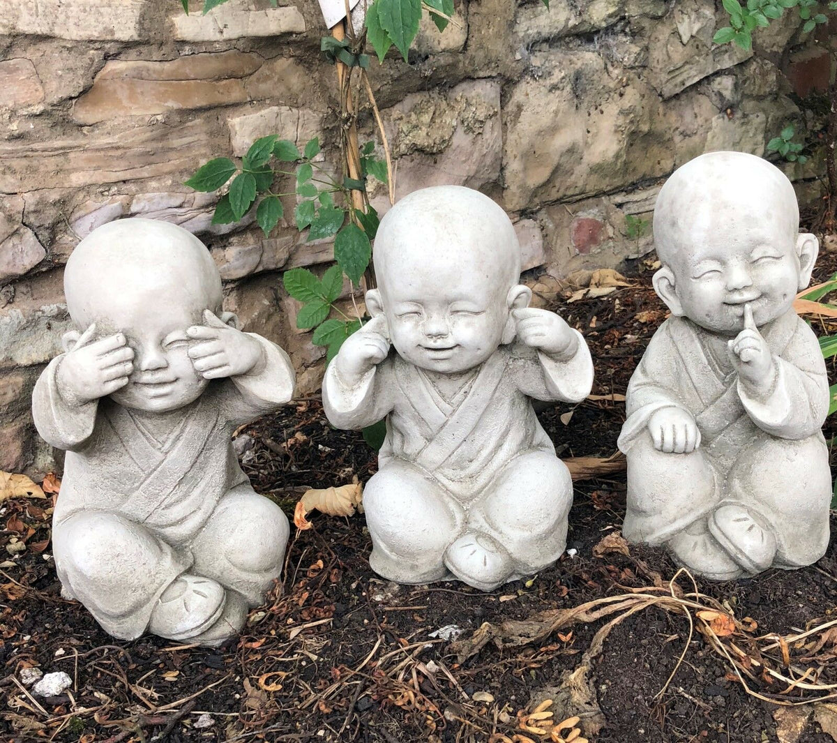 Stone Baby Monk Ornaments trio representing See, Hear, Speak No Evil, placed on soil with leafy backdrop for outdoor decor.