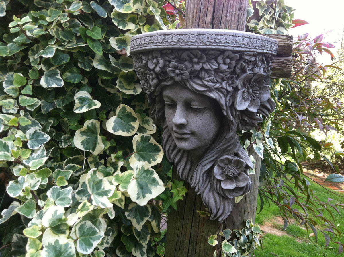 Close-up of Garden Lady Stone Wall Shelf Plaque with floral details, attached to a wooden post among variegated ivy. Elegant garden ornament.