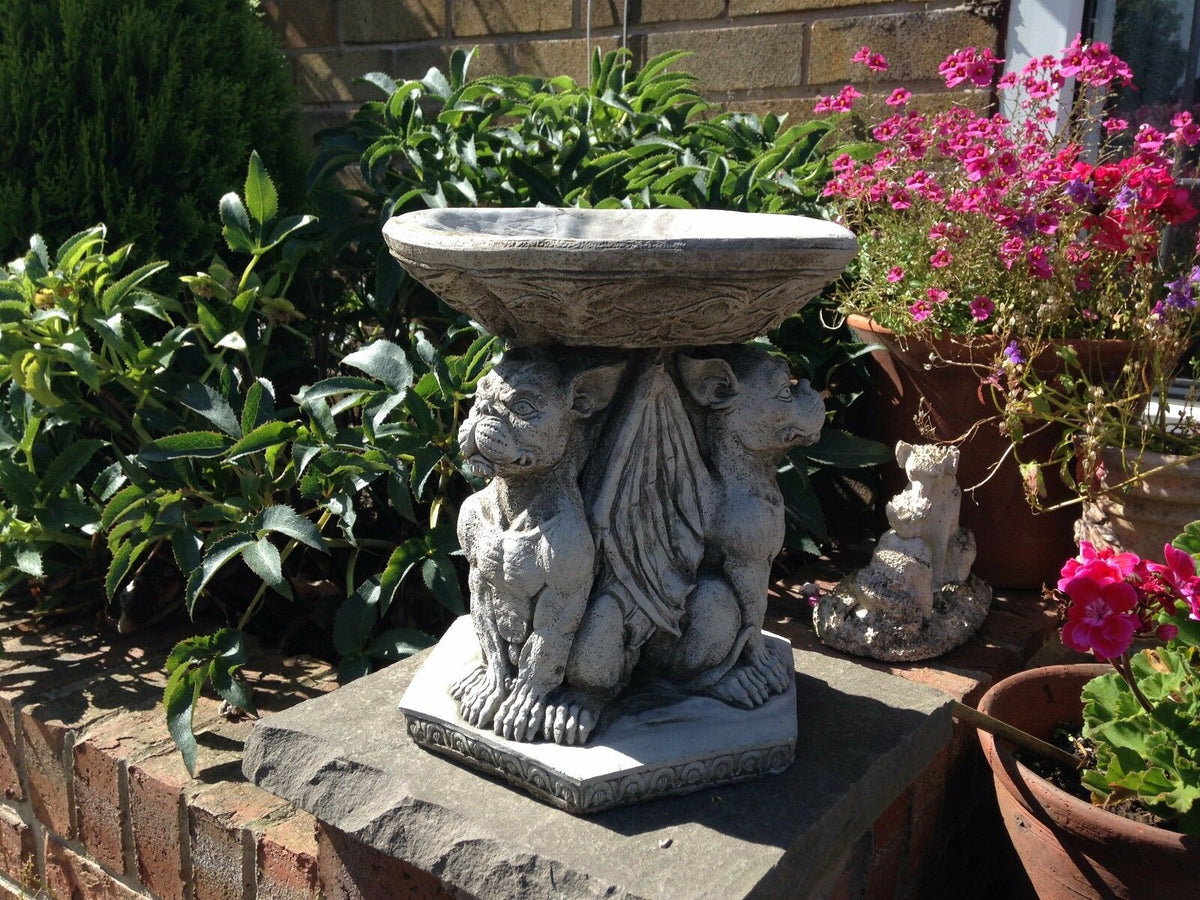 Garden Bird Bath - Stone Gargoyle Design with gothic winged figures, placed on a brick pedestal among potted flowers and greenery.